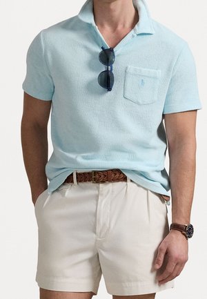 Mężczyzna ubrany w jasnoniebieską koszulkę polo z krótkim rękawem i kieszenią na piersi, granatowe okulary przeciwsłoneczne zwisające z kołnierzyka, beżowe szorty, brązowy pleciony pasek oraz zegarek na nadgarstku.