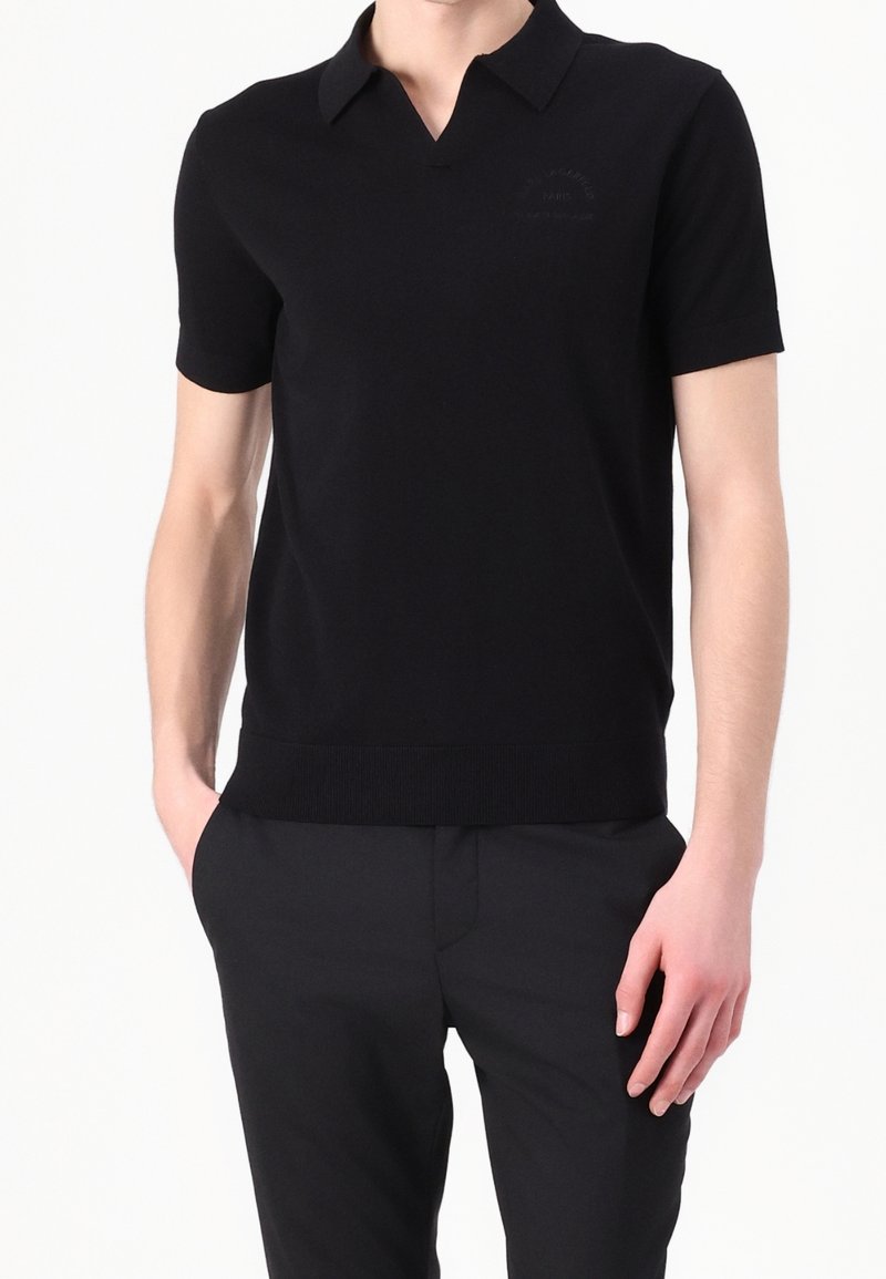 Homme portant un polo noir à manches courtes avec col et un pantalon noir, debout avec une main dans la poche.