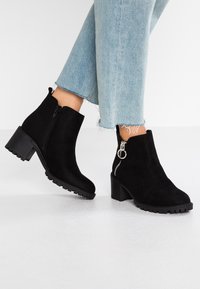 Stivaletti neri in suede con tacco spesso, cerniera laterale e dettaglio decorativo ad anello. La suola testurizzata offre aderenza.