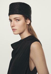 Zwarte gevlochten hoed met een cilindrische vorm en een gestructureerde afwerking. Draag het met een zwarte mouwloze top met zachte draping en een asymmetrische halslijn.