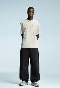 Young man standing, wearing a beige t-shirt with a pink star graphic, black wide-leg pants, silver chain necklace, and gray sneakers.