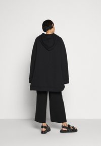 Felpa nera oversize con cappuccio, abbinata a pantaloni neri a gamba larga e sandali neri con dettagli a fibbia. Tessuto dalla texture liscia.
