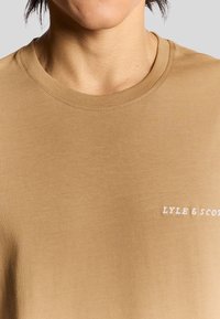 Tan bomull T-shirt med rund halsringning, prydd med vit broderad text "LYLE & SCOTT" på nedre högra sidan. Slät textur.