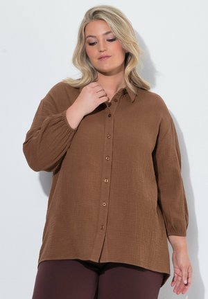 Kvinde med blond hår iført en løs brun skjorte med knapper og lange ærmer samt mørke bukser mod en ensfarvet lys baggrund.
