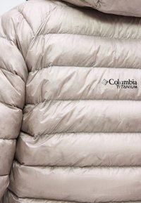 Veste matelassée beige clair avec matelassage horizontal, affichant le logo « Columbia Titanium » sur la poitrine.