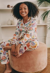Ensemble de pyjama coloré et à motifs avec diverses illustrations de dessins animés, fabriqué en tissu doux. Enfant assis sur un pouf rond rose en peluche.