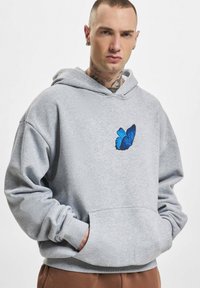 Šedá mikina s kapucí, se vyšitým modrým motýlem na hrudi, uvolněný střih a ribbovanými manžetami. Měkký povrch látky.