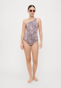 Enosna kopalna obleka z enim ramenom v roza leopardjem vzorcu, oprijetega kroja, gladke teksture s sijočim zaključkom, prikazana na modelu, ki nosi sončna očala.