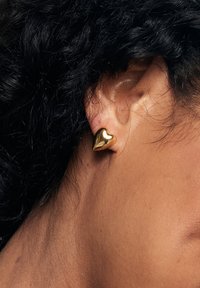 Złote kolczyki w kształcie serca o gładkiej, błyszczącej powierzchni. Noszone na płatku ucha, prezentujące elegancki i minimalistyczny design.