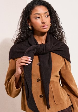 Femme aux cheveux bouclés portant des boucles d'oreilles créoles dorées, une veste en suède marron et une écharpe en tricot noire nouée autour du cou.