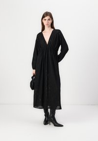 Μαύρο μάξι φόρεμα από δαντέλα με βαθύ V-ντεκολτέ, μακριά μανίκια και συγκεντρωμένη μέση. Φορεμένο με μαύρες μπότες μέχρι το γόνατο με τακούνι και μια μικρή τσάντα.