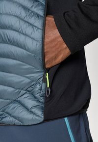 Υβριδικό μπουφάν με κουβερτισμένο μπλε ύφασμα από τη μία πλευρά και μαύρο μαλακό υλικό α�πό την άλλη, διακοσμημένο με φερμουάρ που εκτίθεται και πράσινο τράβηγμα.