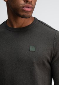 Dunkelgrauer Strickpullover mit Rundhalsausschnitt und einem grünen quadratischen Logopatch auf der linken Seite. Der Stoff hat eine glatte, strukturierte Oberfläche.