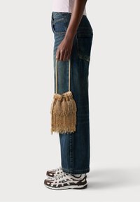 Gold beaded handbag with tassel fringe, gathered top, and a short handle. Worn with blue denim jeans and white with brown sneakers.