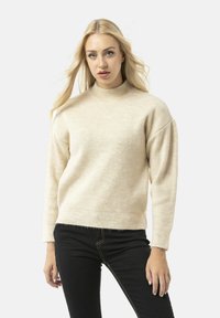 Beige pulloversweater met een ribbeltextuur, hoge nek en verlaagde schouders. Gecombineerd met zwarte jeans met gele stiksels.