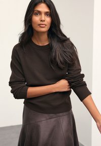 Brown textured sweater with ribbed cuffs and hem, paired with a dark leather skirt. Model poses with arms crossed, minimal background.