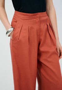 Roodkleurige, geplooide broek met een gestructureerde stof, zijzakken en een hoge taille. Gepaard met een zwarte top en een zilveren armband.