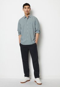 Karierter Button-Up-Hemd in Blau- und Grüntönen, kombiniert mit marineblauen Hosen und weißen Sneakers, vor einem schlichten Hintergrund stehend.
