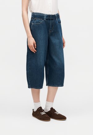 Άτομο που φοράει μπλε σκούρα τζιν σορτς μέσης μήκους, άσπρες κάλτσες, καφέ αθλητικά παπούτσια και ένα φορεμένο μέσα λευκό αμάνικο τοπ.