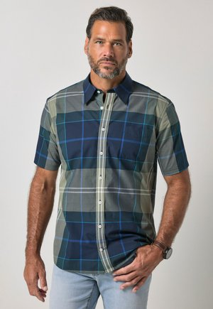Mand med kort mørkt hår og skæg iført en blå og grøn ternet kortærmet skjorte og lyseblå jeans mod en ensfarvet baggrund.