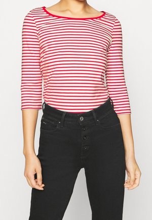 Kobieta ubrana w czerwono-biały top w poziome paski z rękawami do łokcia, włożony w wysokie czarne spodnie z guzikiem na przodzie.