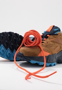 Chaussure de randonnée marron et orange avec une semelle bleu et noir, munie d'un lacet orange vif lâche enroulé dessus et s'étendant devant la chaussure.