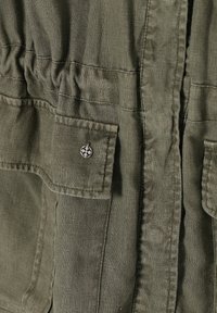 Gros plan d'une veste en tissu vert olive avec des coutures surpiquées, une ceinture froncée et une poche à rabat avec un petit bouton métallique rond.