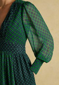 Zielona sukienka w wzory, z przezroczystymi, bufiastymi rękawami w niebieskie kształty diamentów. Gorset ma wyraźnie zaznaczoną talię, a spódnica jest zwiewna.