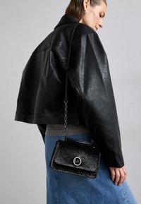 Veste en cuir noir courte avec un sac à main à bandoulière en chaîne ; le sac a une surface brillante, un logo en argent et une forme rectangulaire. Jupe en denim visible.