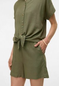 Completo verde oliva a maniche corte con dettaglio annodato in vita, caratterizzato da una chiusura a bottoni e pantaloni shorts larghi. Tessuto di texture liscia.