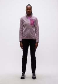 Γυναίκα στέκεται μπροστά, φοράει ανοιχτό μωβ μπουφάν με φερμουάρ, μαύρο παντελόνι και μωβ παπούτσια πεζοπορίας, μπροστά σε απλό λευκό φόντο.