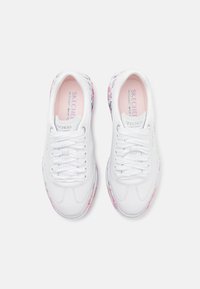 Białe sneakersy Skechers z kwiatowymi akcentami po bokach, zaokrąglonym noskiem, sznurowanym wzorem i wyściełanym kołnierzem. Miękka, teksturowana wkładka.