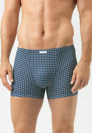 Männlicher Oberkörper, bekleidet mit marineblauen Boxershorts mit weißem Kreis-Muster, stehend mit entspannt an den Seiten hängenden Armen vor einem einfarbigen Hintergrund.