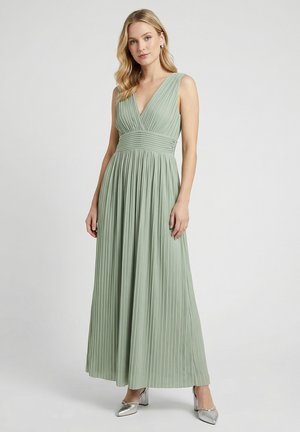 Frau trägt ein ärmelloses, hellgrünes plissiertes Maxikleid mit tiefem V-Ausschnitt und silbernen High Heels, steht vor einem schlichten Hintergrund.