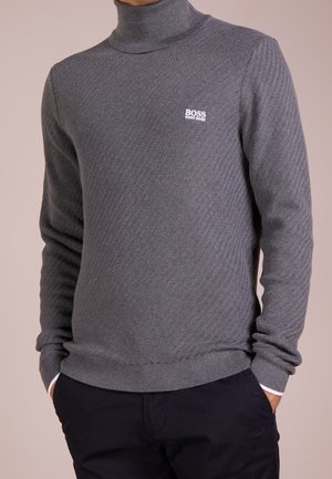 Mann trägt einen grauen Hugo Boss Strick-Rollkragenpullover mit dezentem Strukturmuster und schwarze Hose, Hände in den Taschen.