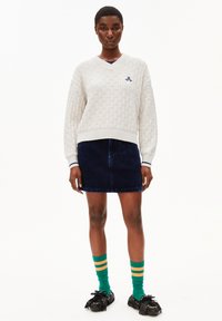 Lichtgrijze gebreide trui met V-hals, marineblauwe corduroy rok, groene sokken met gele strepen en zwarte schoenen met franjes.