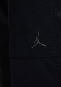 Black fabric pants featuring a stitched gray Jordan logo. The material has a smooth texture with a subtle sheen.