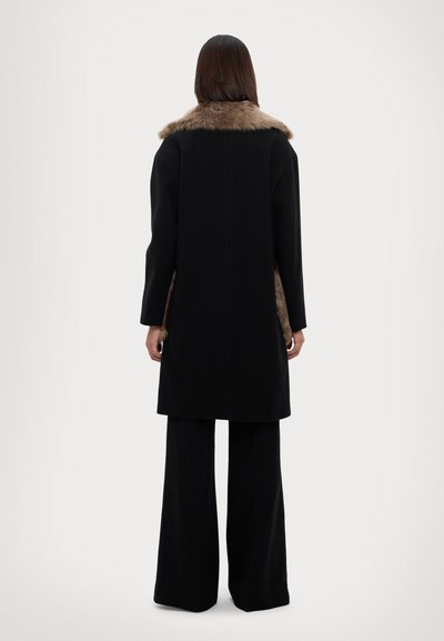 Cappotto di lana nera con colletto in pelliccia sintetica, taglio dritto, maniche lunghe e dettagli laterali, abbinato a pantaloni neri a gamba larga.