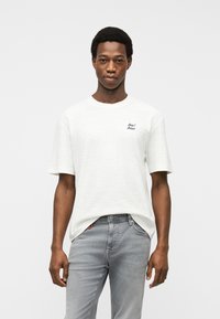 Άνδρας που φοράει λευκό κοντομάνικο T-shirt με μικρό μαύρο κείμενο στο στήθος και ανοιχτό γκρι  τζιν, στέκεται μπροστά σε λευκό απλό φόντο.