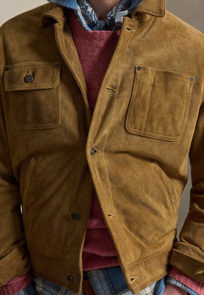 Άνδρας που φοράει μπεζ τζάκετ από σουέντ πάνω από κόκκινο πουλόβερ με πλεκτό σχέδιο και μπλε πουκάμισο με σχέδια, με μπλε κασκόλ με σχέδια γύρω από το λαιμό.