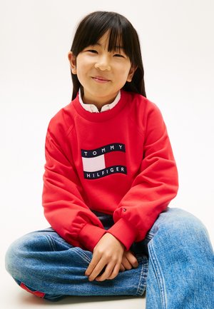 Junges Mädchen sitzt mit überkreuzten Beinen, trägt ein rotes Tommy Hilfiger Sweatshirt und blaue Jeans, lächelt sanft in die Kamera.