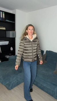 Nafoukaná olivově zelená bunda s vysokým límcem a krémovou podšívkou. Model nosí světle modré džíny a černé boty, chodí po světlé dřevěné podlaze.