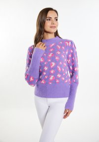 Maglione viola con una texture morbida, caratterizzato da macchie di leopardo rosa e bianche. Collo alto e maniche lunghe con polsi a costine. Indossato con pantaloni bianchi.