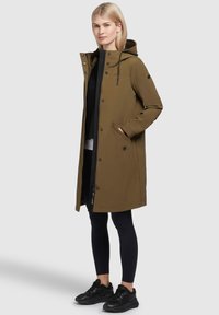 Manteau féminin olive, mi-long, avec capuche, fermeture éclair et boutons-pression, et poches latérales. Porté sur des leggings noirs et des baskets.