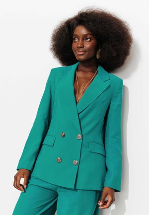 Femme avec une grande coiffure afro portant un blazer croisé vert sarcelle et un pantalon assorti, accessoirisée d'un collier et de boucles d'oreilles en or.