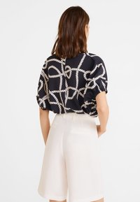 Blusa nera con motivo a corda bianco, vestibilità ampia e maniche arricciate, abbinata a pantaloni corti color crema. La superficie del tessuto è liscia.