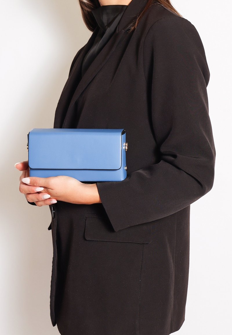 Borsetta blu con una texture liscia, forma rettangolare e chiusura a patta. Tenuta contro un blazer nero. Dettagli in metallo dorato.