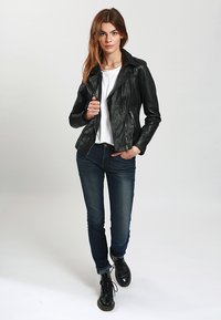 Giubbotto motociclistico in pelle nera con zip frontale, abbinato a una t-shirt bianca e jeans blu scuro. Modello con stivali neri stringati.