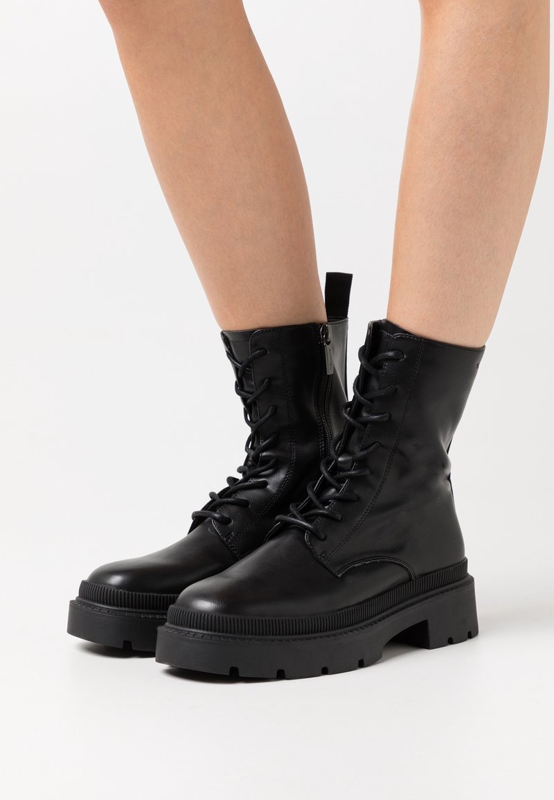 Bottes de combat noires en cuir à lacets avec semelles épaisses portées sur des jambes nues, présentées sur un fond blanc uni.