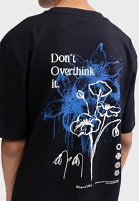 Schwarzes Baumwoll-T-Shirt mit einem blauen Grafikdesign aus Blumen und dem Text "Don't Overthink it", ergänzt durch weiße Linienelemente und Tropfen.